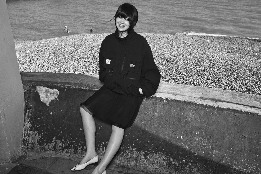 A black-and-white image of a smiling woman sitting on a low concrete ledge by a pebble beach, wearing the JUNYA TACTICAL JACKET and skirt.