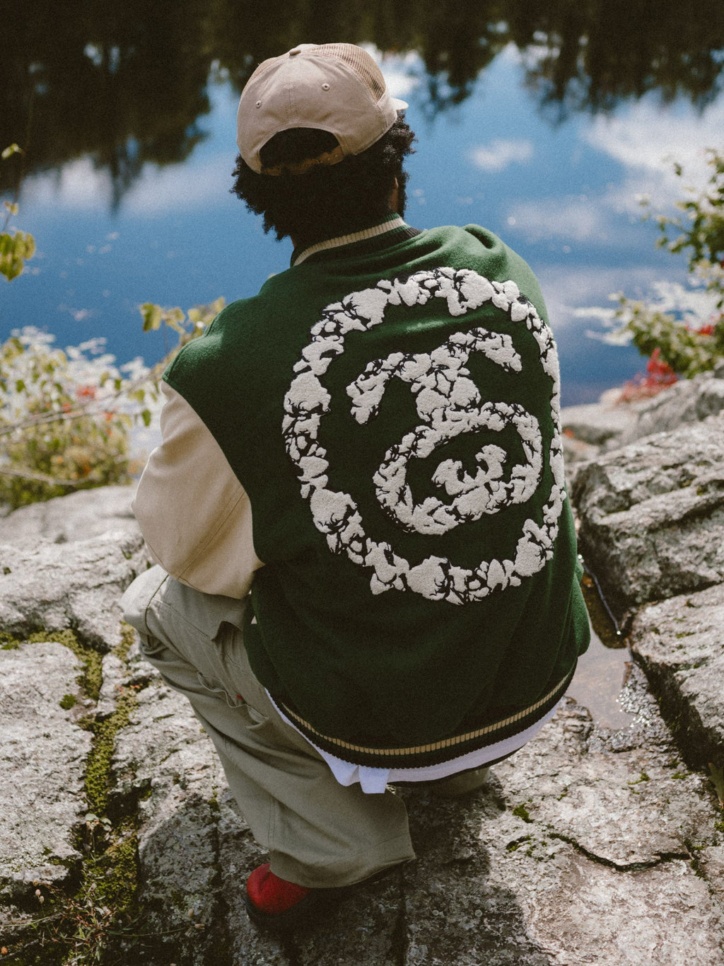 Photo of a model from behind, crouching by a body of water wearing the Denim Tears Varsity Jacket and Denim Tears Cap.