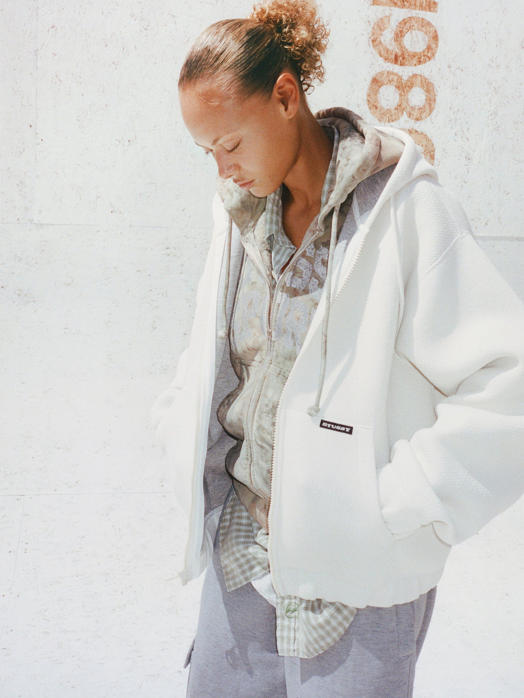 Model wears the Stüssy Bonded Jersey Zip Hoodie in white layered over a plaid shirt with light grey trousers.